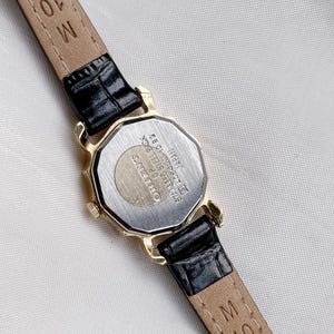 May include: A gold-toned Orient watch with a black leather strap. The watch has an octagonal shape and the back of the watch has the text "STAINLESS STEEL BACK" and "JAPAN". The strap is marked "M 10".