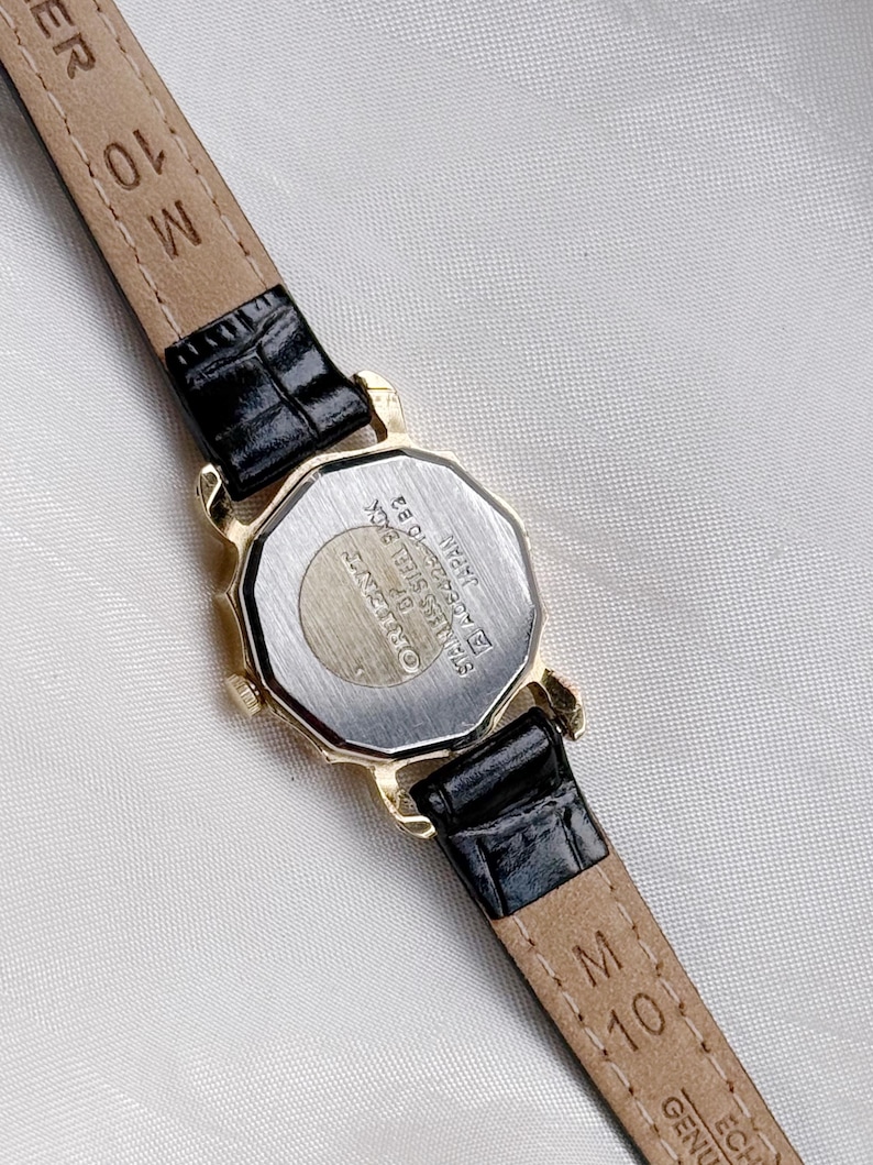 May include: A gold-toned Orient watch with a black leather strap. The watch has an octagonal shape and the back of the watch has the text "STAINLESS STEEL BACK" and "JAPAN". The strap is marked "M 10".