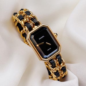 以下が含まれることがあります： CHANELとSWISSの文字が入った八角形の黒い文字盤を持つゴールドトーンの腕時計。時計のブレスレットは、ゴールドトーンの金属チェーンリンクと黒いレザーのストリップが編み込まれており、構造的で層状の外観を作り出しています。