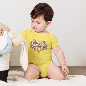 May include: A yellow baby onesie with the word "Schmoopy" in a decorative font, along with the phrase "always & forever." The onesie features a graphic of two birds, hearts, and floral elements. The baby is sitting on a white surface.