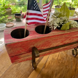 May include: A rustic, red wooden centerpiece with three circular openings, adorned with small American flags and faux greenery. The centerpiece is supported by decorative metal brackets, perfect for patriotic decor.