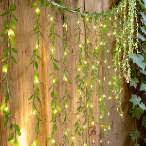 May include: Green leaf string lights with small, warm white LED bulbs hang against a wooden background. The lights are arranged in a cascading pattern, creating a natural, decorative effect. The leaves are a vibrant green, and the lights are evenly spaced.