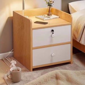Puede incluir: Una mesita de noche de madera clara con dos cajones blancos y una superficie superior. La parte superior tiene un libro, un teléfono inteligente y un jarrón con flores. Una taza de café está en el suelo frente a la mesita de noche.