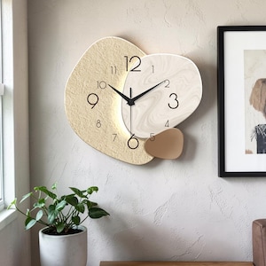 Puede incluir: Un reloj de pared asimétrico con una sección izquierda de color beige texturizado y una sección derecha de color crema con efecto mármol liso. La esfera muestra números arábigos negros del 1 al 12, manecillas negras finas y un detalle dorado.