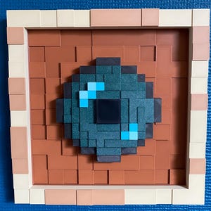 May include: A pixel art piece with a teal and black circular design and light blue highlights, set against a brown brick-effect background. The artwork is framed with a beige and light brown border, creating a 3D effect.