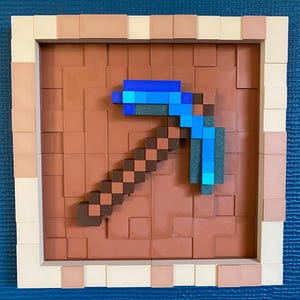 Puede incluir: Una pieza de arte pixelado enmarcada con un diseño de pico. El pico está representado en tonos marrones y azules, sobre un fondo marrón texturizado. El marco es una combinación de beige y marrón claro. La obra de arte es un guiño a la estética de los videojuegos.