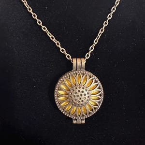 May include: A gold-colored sunflower pendant necklace. The pendant features a detailed sunflower design with a yellow center and petal accents. The necklace has a delicate chain and is set against a dark background.