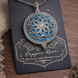 May include: A silver-toned pendant necklace with a circular, ornate design. The pendant features a bright blue enamel background with a swirling pattern and clear crystal accents. The necklace is displayed on a black card with the text "Copper Rose Designs and Engraving."