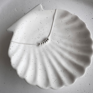 Könnte beinhalten: Eine zarte Silberkette mit einer Reihe kleiner, runder Perlen. Die Kette liegt auf einer weißen, muschelförmigen Schale mit gesprenkelten Details. Der Hintergrund ist eine neutrale, helle Farbe.
