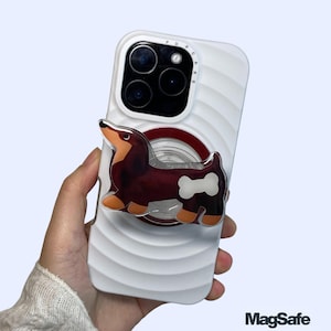 May include: White phone case with a dachshund dog MagSafe phone stand and grip. The dog is brown, black, and orange with a white bone detail. The phone case has a ridged design.