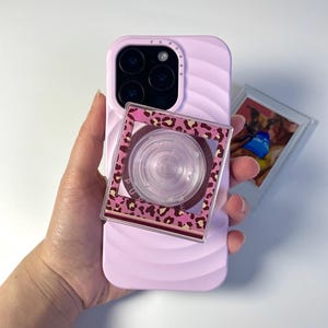 Op de afbeelding: Lila telefoonhoesje met een pop-up grip met luipaardprint. De hoes heeft een gegolfde textuur. Een kleine foto is zichtbaar naast de telefoon.