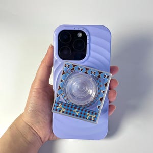Może przedstawiać: Lawendowe etui na telefon z teksturowanym tyłem i wzorzystym uchwytem na telefon. Uchwyt na telefon ma przezroczysty środek i kwadratowy wzór z niebiesko-brązową kratą i żółtymi kokardkami.