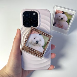 May include: A light pink phone case with a white dog photo and a brown and white patterned frame. The phone case has a wavy design and the word "PUPCASE" printed on the top. Another photo of the dog is in a clear frame.