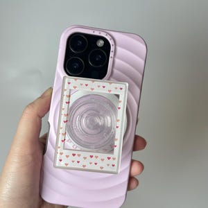 Puede incluir: Una funda de teléfono rosa claro con un diseño de remolino, que sostiene un agarre de teléfono transparente con un marco con estampado de corazones. La funda del teléfono tiene una protuberancia de la cámara con múltiples lentes.
