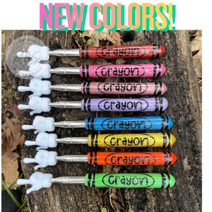 May include: A collection of colorful crayon-shaped bottle stoppers. Each stopper is a different color, including red, pink, purple, blue, yellow, orange, and green, with the word "Crayon" printed on them. The stoppers have a white, cartoon-style hand design.