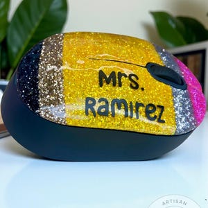 May include: A black wireless computer mouse with a glittery top. The top features gold, silver, and pink glitter stripes, with the name "Mrs. Ramirez" in black lettering. The mouse has a black base and a circular logo that says "Artisan Glitter Studio".