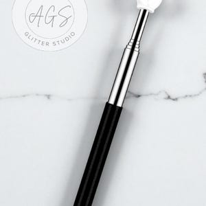 May include: A silver and black retractable pointer with a white hand-shaped tip, pointing upwards. The pointer has a black, textured grip and the text "ARTISAN AGS GLITTER STUDIO" in a circular logo.
