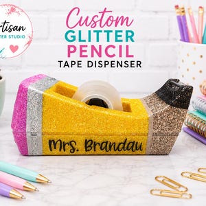 May include: A desk tape dispenser shaped like a pencil, covered in pink, silver, yellow, and black glitter. The yellow section features the black text Mrs. Brandau. It holds a roll of clear tape and sits on a white surface with office supplies.