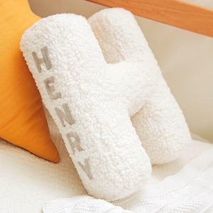 May include: A white, fluffy, letter "H" shaped pillow with the name "HENRY" embroidered in gray. The pillow is made of a soft, textured material and rests on a white blanket. An orange pillow is visible in the background.