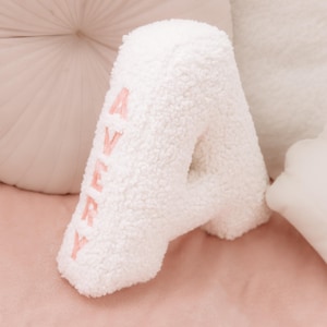 May include: A white, fluffy, letter "A" pillow with the name "Avery" embroidered in pink. The pillow is made of a soft, textured material and is surrounded by other pillows on a pink surface.