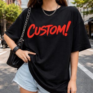 May include: Black t-shirt with the word "CUSTOM!" in red, brush-style lettering. The shirt is worn with light blue denim shorts and a black purse with a silver chain strap. A silver chain necklace is also visible.