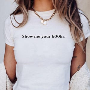 May include: White crop top with the text "Show me your books." The shirt is short-sleeved and has a round neckline. The model is wearing a gold chain necklace and a cream-colored cardigan.
