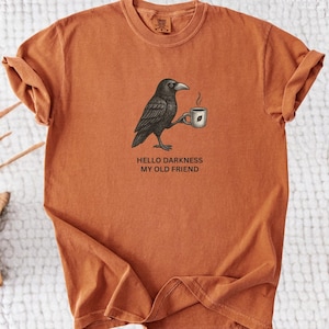 May include: Burnt orange t-shirt featuring a black crow holding a coffee cup. The graphic includes the text "Hello Darkness My Old Friend". The shirt has rolled-up sleeves and a relaxed fit. The shirt is made of a soft, comfortable material.