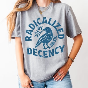 Könnte beinhalten: Graues T-Shirt mit dem Text "RADICALIZED BY BASIC DECENCY" in Blau, mit einem blauen Vogel, der einen Zweig hält. Das Shirt ist aus Athletic Heather-Material.