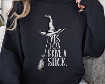 May include: Black sweatshirt featuring a white witch's hat and broom graphic, with the text "YES I CAN DRIVE A STICK." The sweatshirt has a crew neck design.