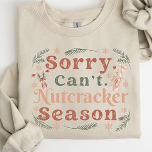 May include: A sand-colored sweatshirt with the text "Sorry. Can't. Nutcracker Season" in a festive design. The design includes candy canes, ballet shoes, snowflakes, and sprigs of greenery.