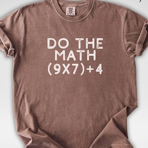 May include: Espresso-colored t-shirt with the phrase "DO THE MATH (9X7)+4" printed in white. The shirt has a crew neck and short sleeves, with the sleeves rolled up. The shirt is made of a soft, comfortable material.