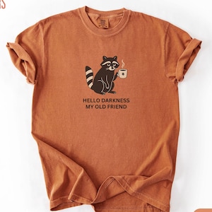 Könnte beinhalten: Ein rostfarbenes Kurzarm-T-Shirt mit einer Grafik eines Waschbären, der eine dampfende Tasse hält. Unter dem Waschbären steht der Text HELLO DARKNESS MY OLD FRIEND. Das T-Shirt hat einen Rundhalsausschnitt und eine lockere Passform.