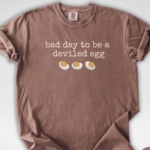 May include: Espresso-colored t-shirt with the phrase "bad day to be a deviled egg" and three deviled egg illustrations. The shirt has a crew neck and short sleeves. The Aurose Studio logo is in the bottom left corner.