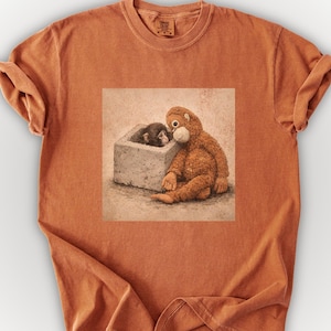 May include: Burnt orange t-shirt featuring a graphic of two orangutans. One orangutan is sitting next to a small concrete box, while the other is inside the box. The image has a vintage, hand-drawn style.