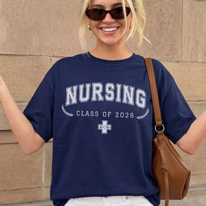 May include: A navy blue short-sleeved t-shirt featuring the white text NURSING CLASS OF 2026 with a small medical cross icon containing a heart and heartbeat line. The shirt is paired with white pants and a brown shoulder bag.