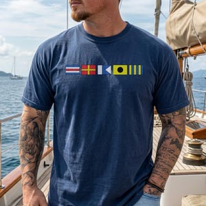 May include: A navy blue t-shirt featuring a series of nautical signal flags across the chest. The flags are in various colors, including red, yellow, and blue. The shirt is made of a soft material and is being worn on a boat.