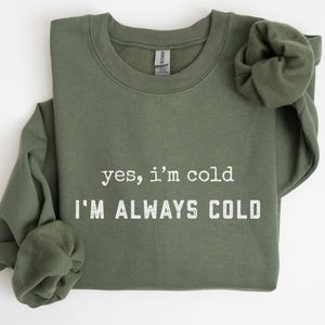 Może przedstawiać: Oliwkowa bluza z białym napisem "yes, I'm cold I'M ALWAYS COLD". Rękawy są podwinięte. Widoczna jest metka marki Gildan. Bluza jest złożona.