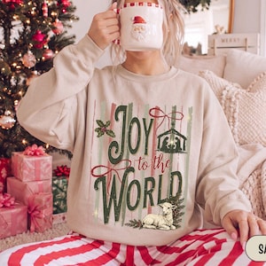 May include: A sand-colored crewneck sweatshirt featuring a graphic print with the text "Joy to the WORLD". The design includes a nativity scene, a sprig of holly, and a lamb illustration, all set against vertical stripes in muted green and red tones.
