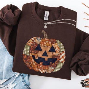 May include: A dark chocolate-coloured sweatshirt with a patchwork pumpkin design. The pumpkin has a smiling face and stem, made from various autumn-themed fabrics. The sweatshirt has a crew neck and long sleeves. The text "Dark Chocolate" is in the upper right corner.
