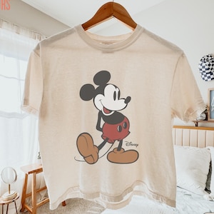 May include: Ivory-colored t-shirt featuring a classic Mickey Mouse graphic. The cartoon character is depicted in a walking pose, wearing red shorts and brown shoes. The shirt has the text "Comfort Colors" on the upper left.