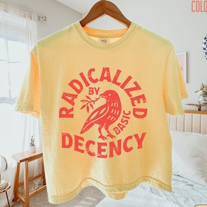 Może przedstawiać: Bladożółty t-shirt z krótkim rękawem z nadrukiem w kolorze wyblakłej czerwieni przedstawiającym ptaka trzymającego gałązkę. Napis RADICALIZED BY BASIC DECENCY ułożony jest w okrąg wokół ptaka. Koszulka ma okrągły dekolt i luźny krój.