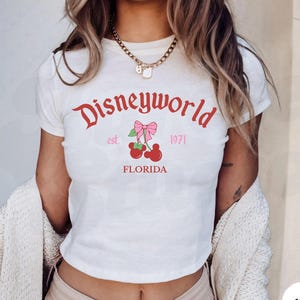May include: White crop top with the text "Disneyworld" in red script, featuring a cherry design with a pink bow. The text "est. 1971 FLORIDA" is also printed on the shirt. The shirt is a casual style.