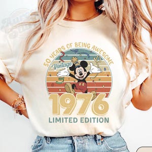 Puede incluir: Camiseta de color marfil con un dise&ntilde;o vintage de Mickey Mouse. El gr&aacute;fico incluye el texto "50 Years of Being Awesome", "Vintage", "1976" y "Limited Edition". Mickey se muestra celebrando dentro de un dise&ntilde;o circular.