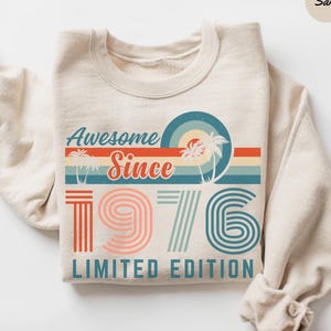May include: Sand-colored sweatshirt with a retro design. The graphic reads "Awesome Since 1976 Limited Edition" in a vintage font, with palm trees and a sunset design. The numbers are in a retro font with orange, pink, and blue colors.