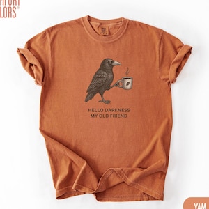 Könnte beinhalten: Ein rostorangefarbenes Kurzarm-T-Shirt mit einer schwarzen Grafik eines Raben, der eine dampfende Kaffeetasse hält. Unter dem Raben steht der Text HELLO DARKNESS MY OLD FRIEND. Das T-Shirt hat einen Rundhalsausschnitt und eine lockere Passform.