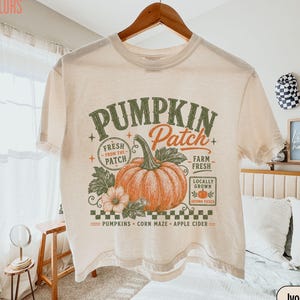 Pode incluir: Uma t-shirt de manga curta na cor marfim com uma estampa gráfica com o texto "PUMPKIN PATCH" em verde e laranja. O design inclui uma abóbora laranja grande, folhas verdes, uma borda xadrez e "PUMPKINS CORN MAZE APPLE CIDER".