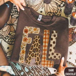 Op de afbeelding: Een donker chocoladebruin sweatshirt met ronde hals en het woord FALL in grote blokletters. Elke letter heeft een eigen patroon: de F is bloemrijk, de A is luipaardprint, de L is oranje ruitjes en de L is bruin met stippen.