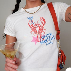 May include: White t-shirt featuring a red and blue lobster graphic holding a martini glass, with the text "Lobster Martini Society" in blue. A pink star is behind the lobster. An orange purse hangs from the shoulder.