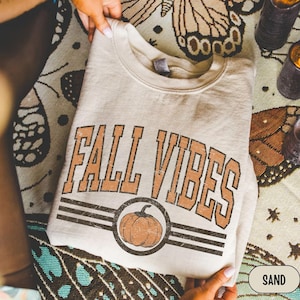 Könnte beinhalten: Ein beigefarbenes Sweatshirt mit Rundhalsausschnitt und dem Schriftzug FALL VIBES in verwaschener brauner Schrift. Unter dem Text befindet sich eine kreisförmige Grafik mit einer braunen Kürbisillustration zwischen drei schwarzen Linien.