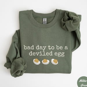 May include: Military green sweatshirt with the phrase "bad day to be a deviled egg" in white text. Below the text are three cartoon deviled eggs. The sweatshirt is a solid color and has a crew neck.